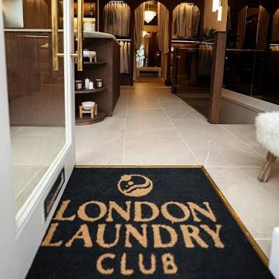 A dark navy doormat with gold lettering reading 'LONDON LAUNDRY CLUB' and a circular club logo, placed at the entrance of a modern interior with tiled floors and warm wood accents.